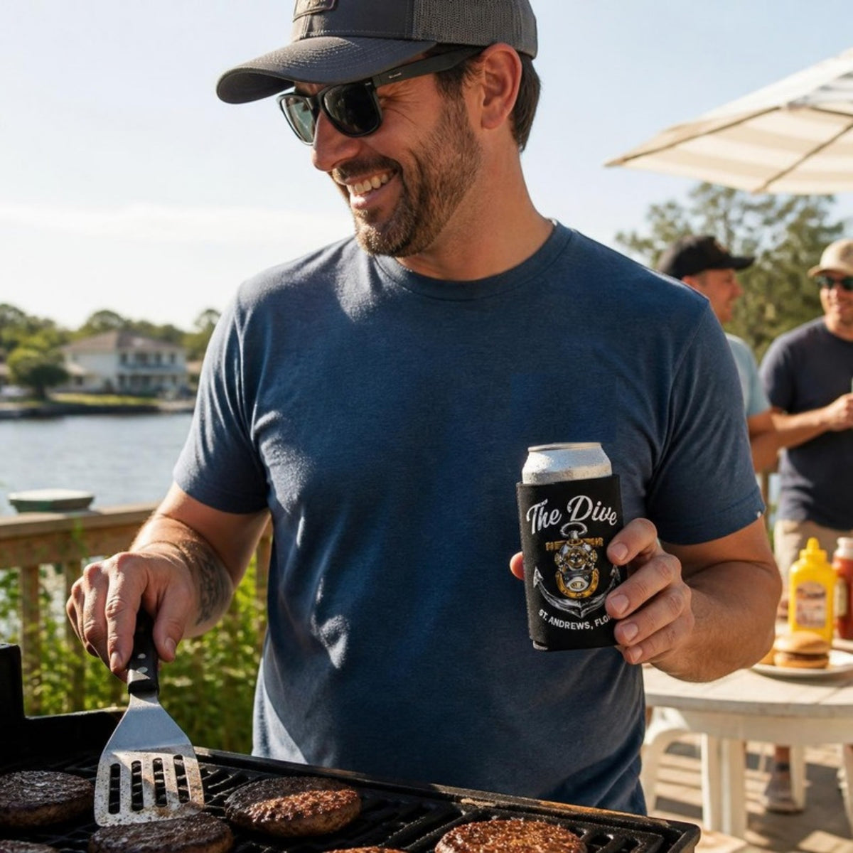 Man grilling by the water with The Dive can koozie
